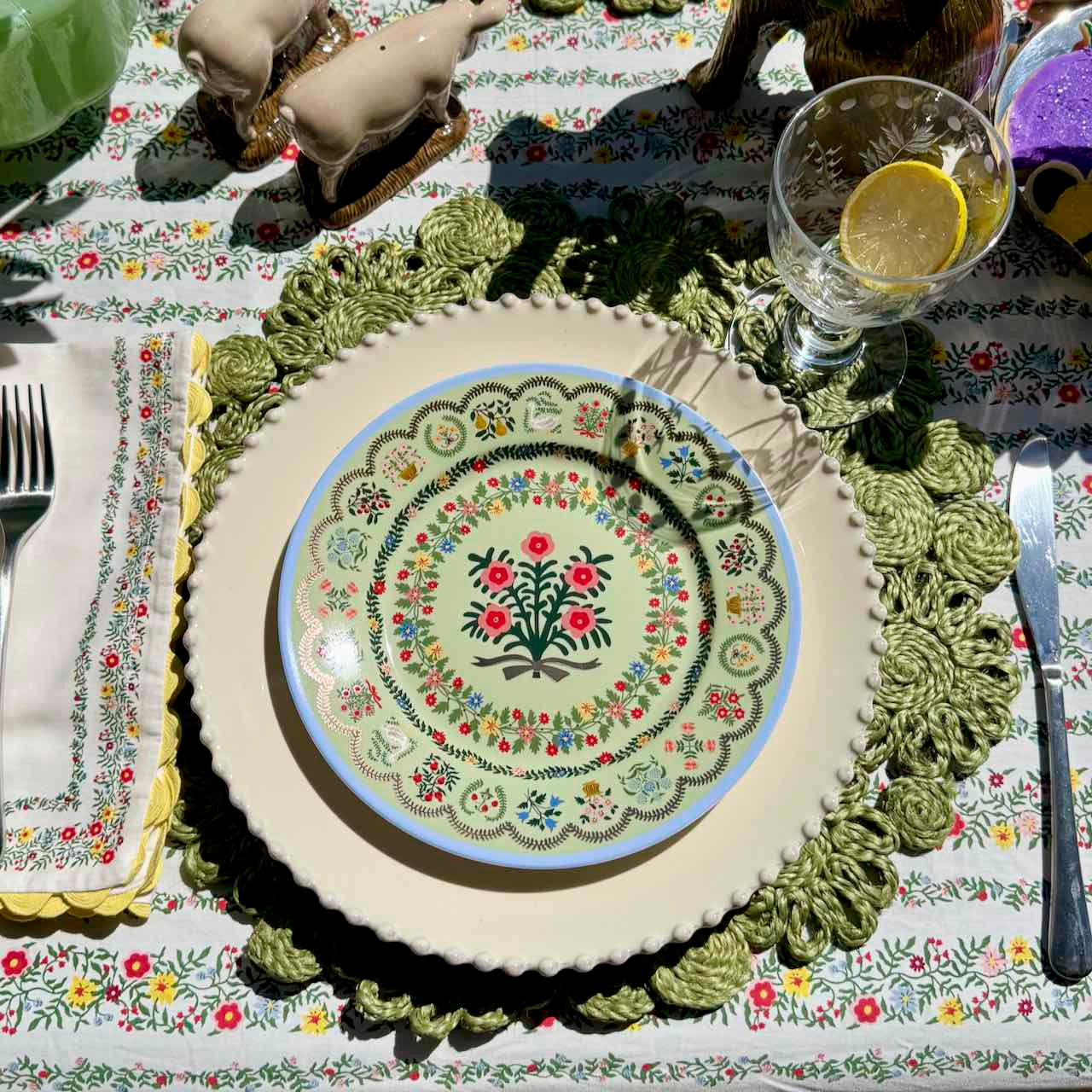 Decorative table setting with a floral plate on a green lace doily, surrounded by cutlery and glasses.