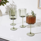 Four fluted stemware glasses in a translucent sage green glass, arranged on a white countertop with one glass containing an iced iced coffee garnish.