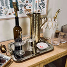 A styled bar setup on a wooden console table featuring a hammered gold metal pitcher, a silver tray, a dark wine bottle with a gold owl decanter, two clear coupe glasses, and a white heart-shaped holder with gold accents.