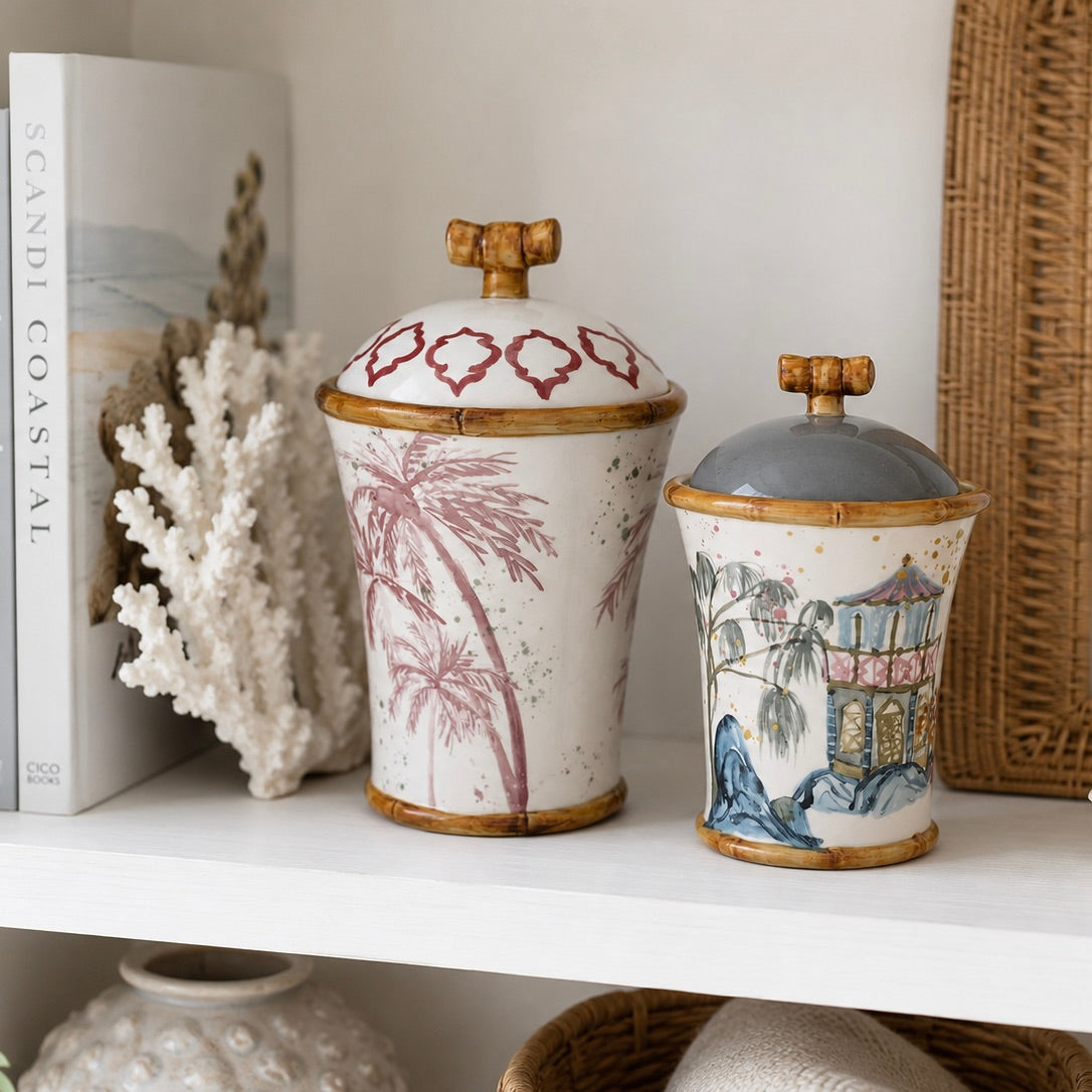 Two decorative lidded ceramic jars with bamboo-style rims and handles, displayed on a white shelf. The larger jar has a white body with pink palm tree illustrations and a red geometric pattern on the lid.