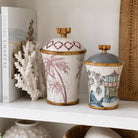 Two decorative lidded ceramic jars with bamboo-style rims and handles, displayed on a white shelf. The larger jar has a white body with pink palm tree illustrations and a red geometric pattern on the lid.
