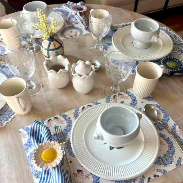 Dining table set with white ceramic dishes, glassware, and decorative elements on a patterned tablecloth.