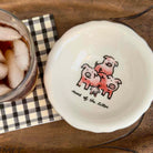 A white ceramic dish with a scalloped edge, featuring a cute illustration of four pink pigs and the text 'smile of the kitten' printed on it. The dish is shown on a wooden table next to a glass of iced cola on a black-and-white checkered napkin.