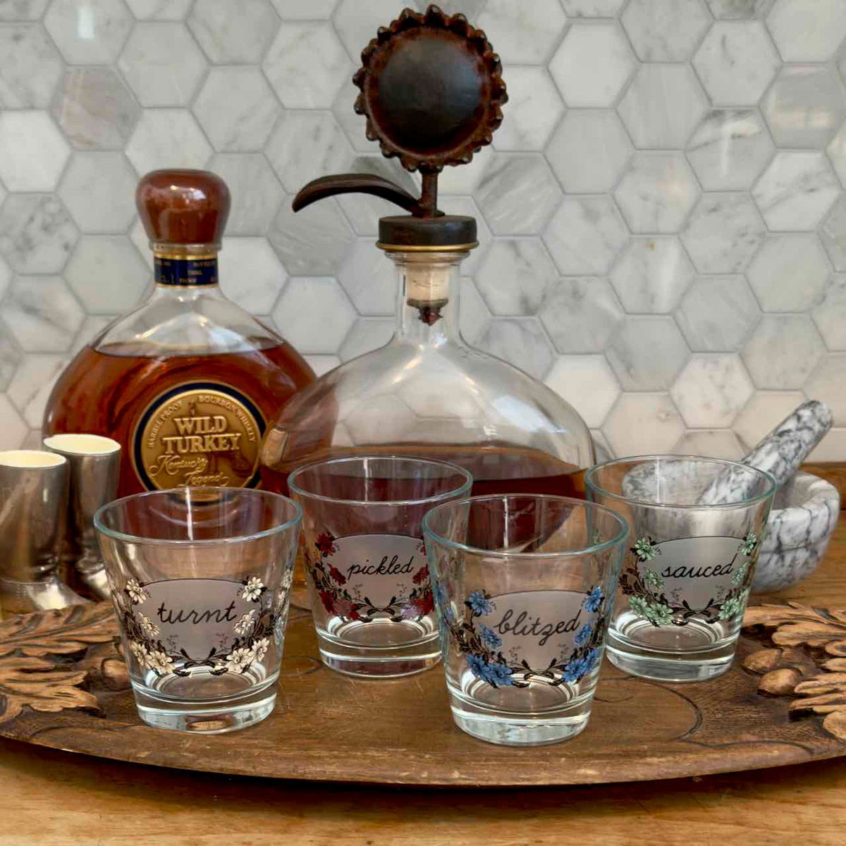 Four clear glass shot glasses with floral designs and handwritten labels reading 'turnt', 'pickled', 'blazed', and 'saucy', arranged on a wooden tray beside a whiskey bottle and decanter in a kitchen setting.
