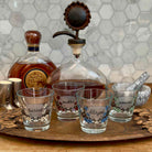 Four clear glass shot glasses with floral designs and handwritten labels reading 'turnt', 'pickled', 'blazed', and 'saucy', arranged on a wooden tray beside a whiskey bottle and decanter in a kitchen setting.