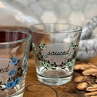 Two clear glass shot glasses with floral designs and the word 'sauced' on the front, displayed on a wooden surface with dried flowers and a blurred background.