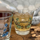A clear glass tumbler with a floral design and the word 'sauced' printed on it, filled with an amber drink and ice, set on a wooden surface. Another matching glass is partially visible at the left edge, and nuts appear at the lower right.