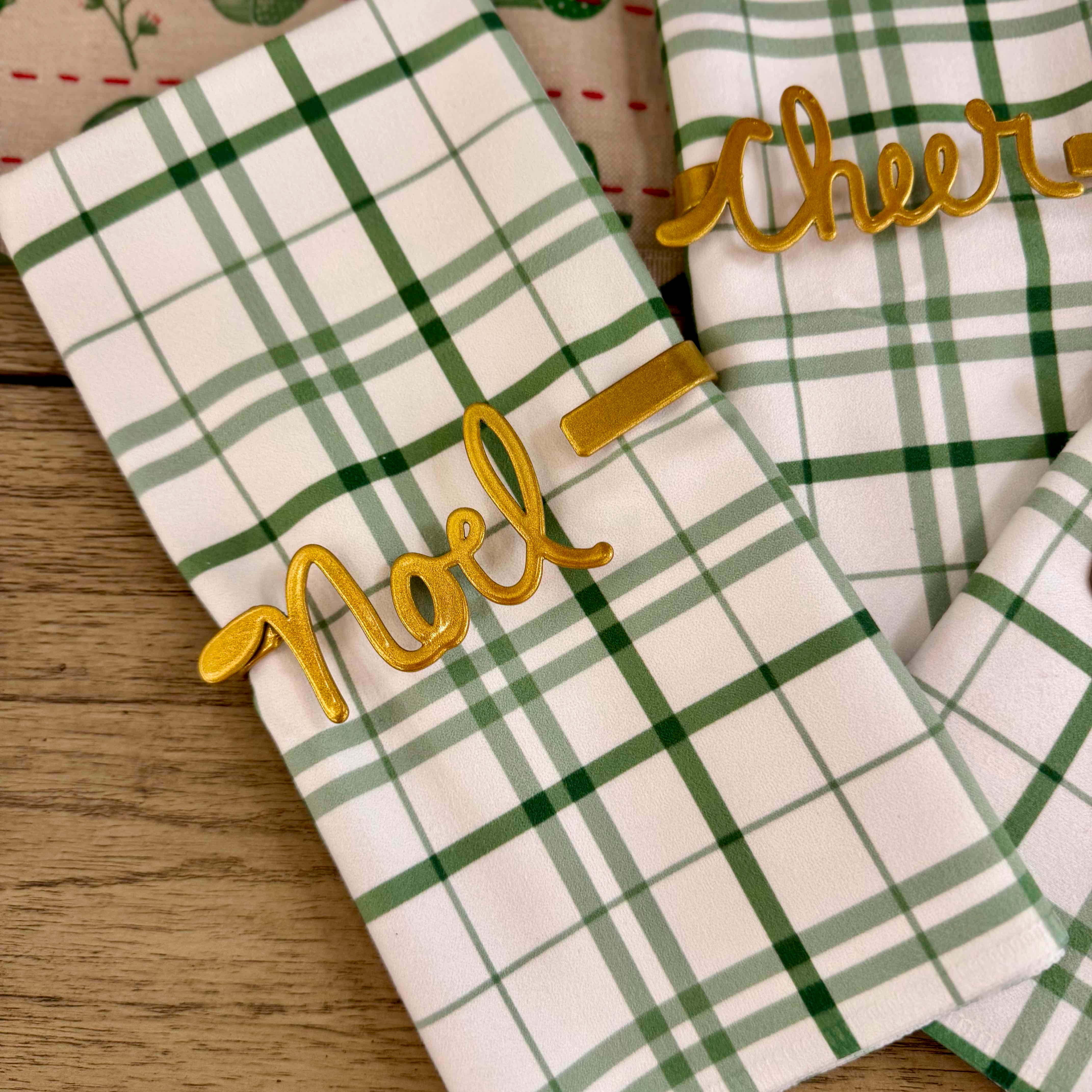 Green and white plaid fabric with gold 'Noel' and 'Cheer' text on a wooden surface