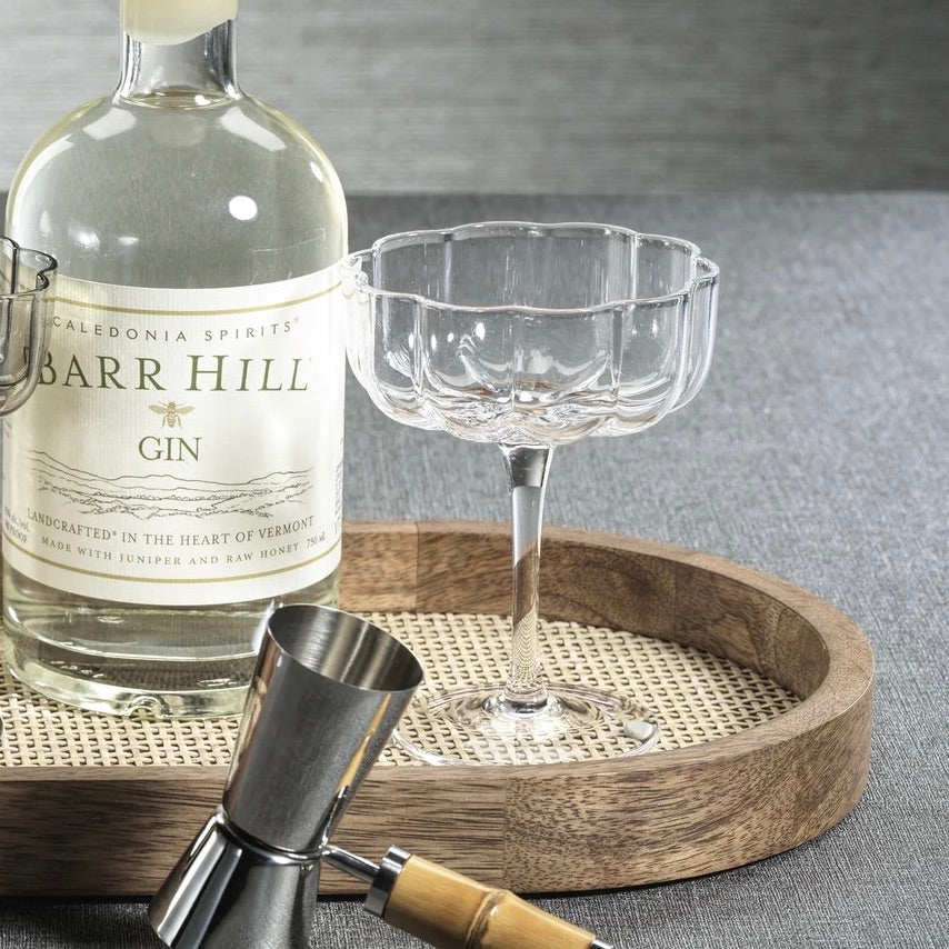 A clear stemmed coupe-style glass and a bottle of Barr Hill Gin are arranged on a round wooden tray with a woven center, alongside metal jigger tools on a gray fabric surface.