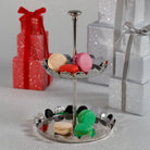 Two-tiered silver stand with colorful macarons against a glittery background with gift boxes.