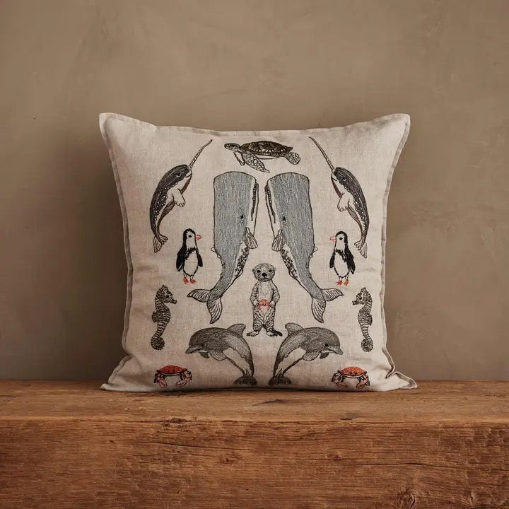 A square decorative throw pillow with an embroidered marine life pattern, resting on a rustic wooden surface against a textured beige wall.