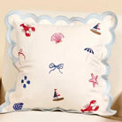 A square decorative throw pillow with a scalloped light blue border and embroidered nautical motifs including red lobsters, a pink seashell, blue umbrellas, and bows on an off-white background.