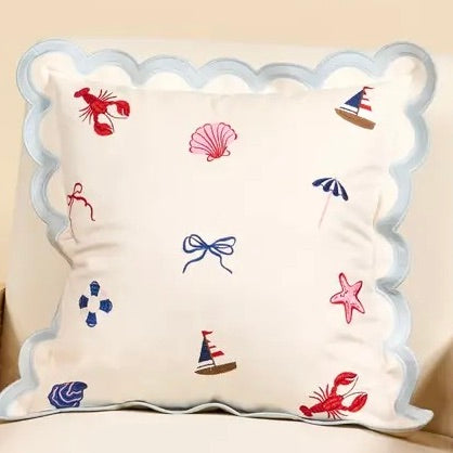 A square decorative throw pillow with a scalloped light blue border and embroidered nautical motifs including red lobsters, a pink seashell, blue umbrellas, and bows on an off-white background.
