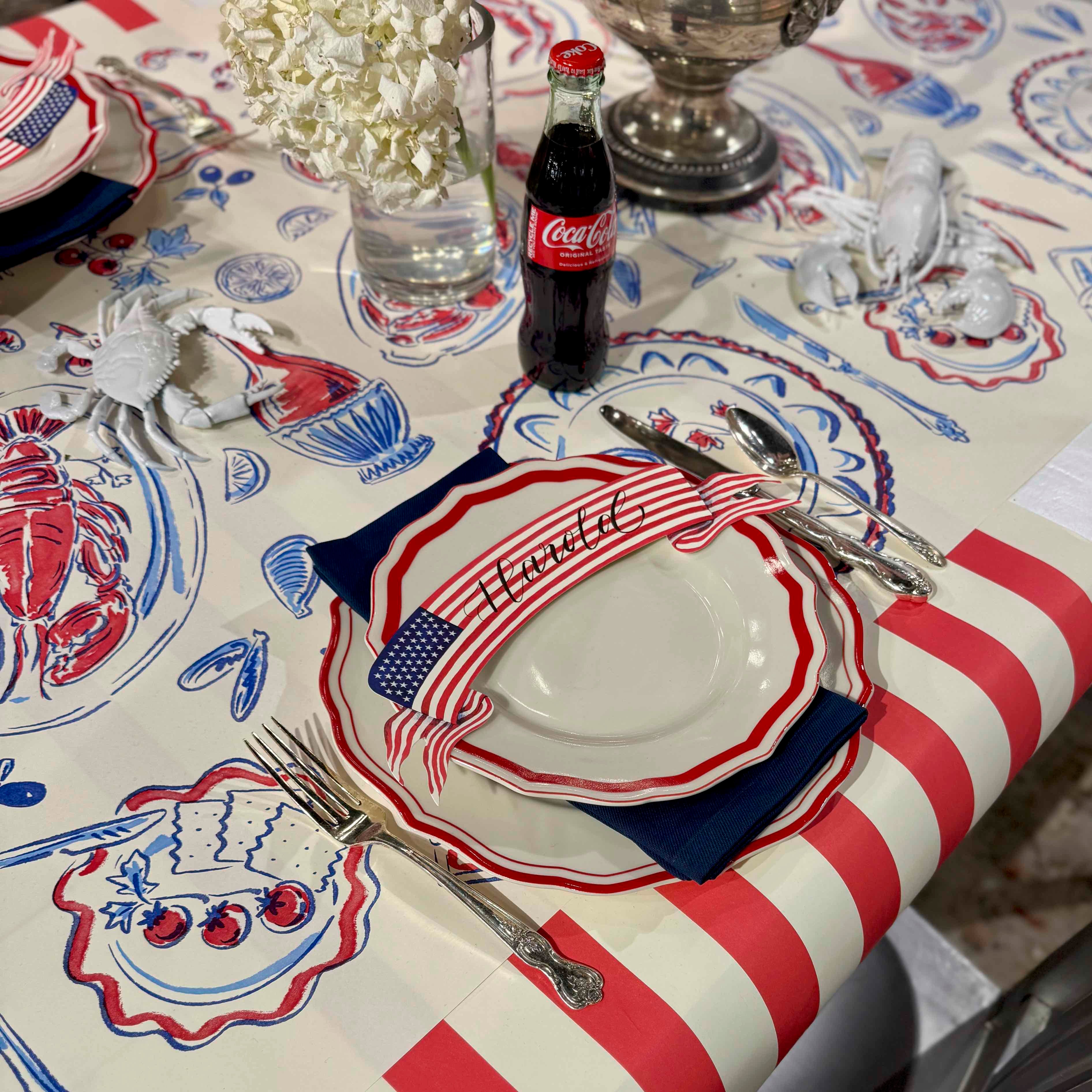 Table setting with a Coca-Cola bottle on a tablecloth featuring American flag and lobster designs.