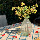 A clear textured glass vase filled with artificial green berries and leaves, placed on a table with a floral tablecloth in an outdoor garden setting.