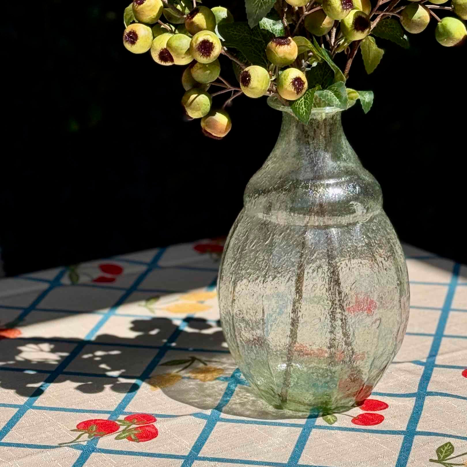 A textured, clear glass vase filled with artificial green leaves and yellow-green berries, set on a patterned tablecloth in natural sunlight.
