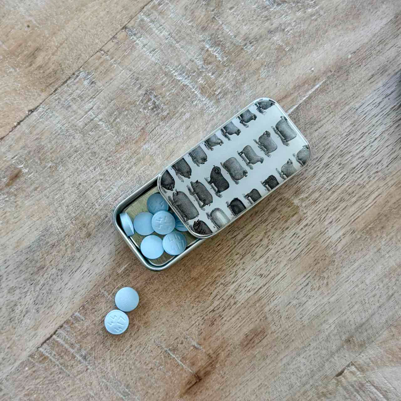 A small silver metal pill organizer tin with a hinged lid, shown open on a wooden surface. Several light blue round tablets are visible inside the compartments, and two more tablets lie outside the tin on the wood.