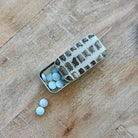 A small silver metal pill organizer tin with a hinged lid, shown open on a wooden surface. Several light blue round tablets are visible inside the compartments, and two more tablets lie outside the tin on the wood.