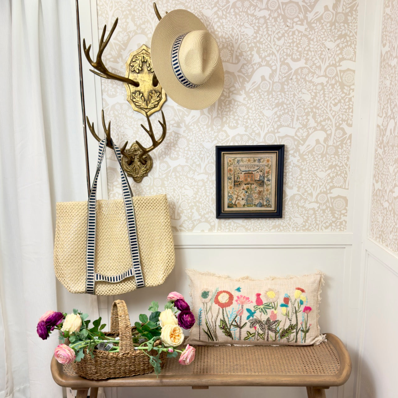 Decorative entryway with a bench, floral pillow, straw bag, and framed picture on a patterned wall.