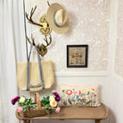 Decorative entryway with a bench, floral pillow, straw bag, and framed picture on a patterned wall.