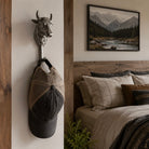 A silver metal wall hook shaped like a bull's head hangs on a white wall in a rustic bedroom setting, holding a beige and black baseball cap. A wooden bed frame, neutral bedding, and a framed mountain landscape print are visible in the background.