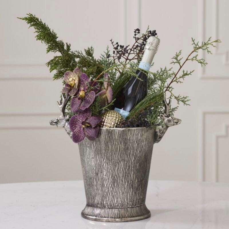 A textured silver champagne bucket filled with green pine branches, purple orchids, a small gold pinecone, and a dark bottle with a pale blue foil, set on a white surface against a cream paneled wall.