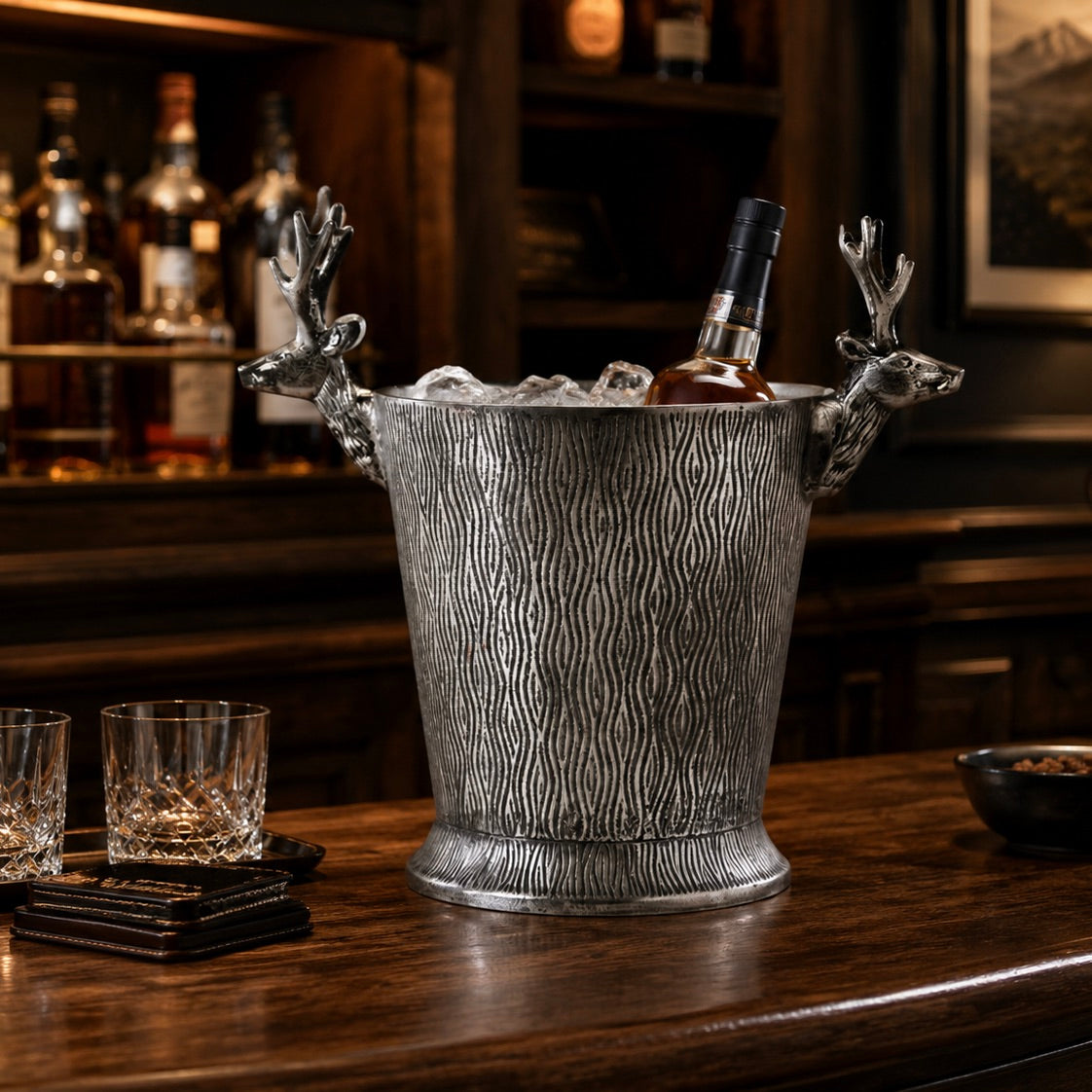 A silver metal whiskey bucket with textured wood grain detailing and decorative deer head handles, filled with ice on a wooden bar counter in a bar setting.