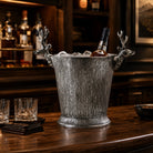 A silver metal whiskey bucket with textured wood grain detailing and decorative deer head handles, filled with ice on a wooden bar counter in a bar setting.