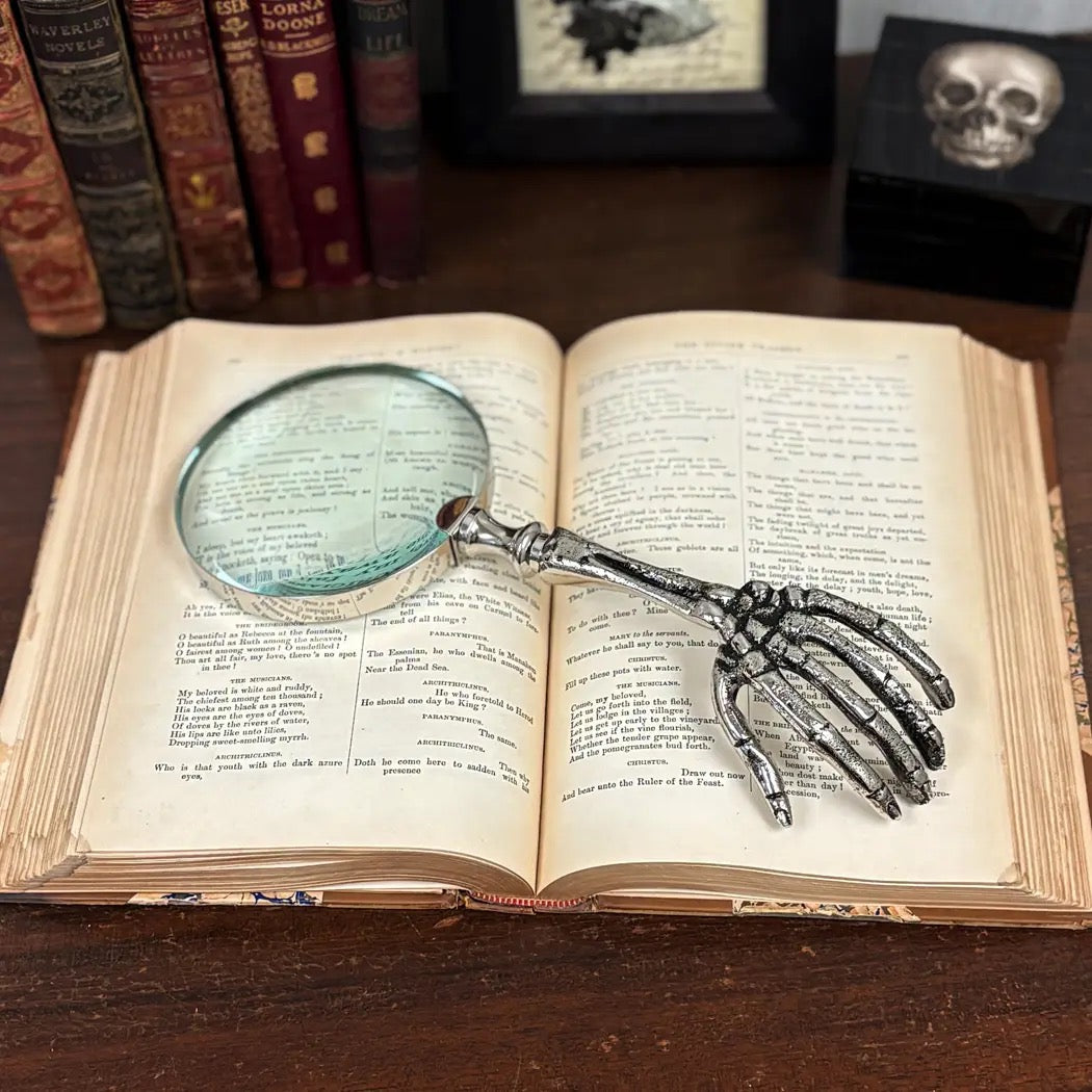 A skeleton hand-shaped magnifying glass resting on an open vintage book on a wooden table, with old books and a skull box blurred in the background.