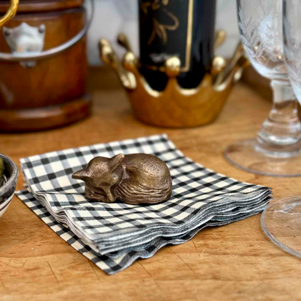 A small metallic sleeping cat figurine sits on a black-and-white checkered napkin on a wooden tabletop, with blurred wine glasses and a bottle in the background.