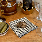A small metallic sleeping dog figurine rests on a black-and-white gingham napkin on a wooden table, surrounded by dining props including pickles, a bottle, and glassware.