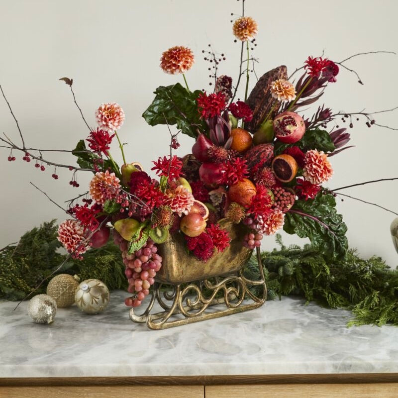Decorative floral arrangement in a gold sleigh on a marble surface with greenery in the background.