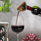 Red wine being poured into a glass with a decorative bottle stopper, grapes, and a plant in the background.