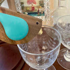 Bird-shaped object with a turquoise body and wooden wings near a glass on a table.