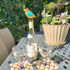 Wine bottle with a decorative bird on top, two glasses, and a potted plant on a table outdoors.