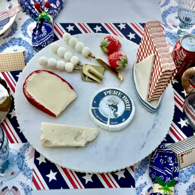 Marble cheese board with assorted cheeses, strawberries, and a can of Coca-Cola on a patriotic tablecloth.