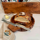 Loaf of bread on a wooden cutting board with a knife, on a tablecloth.