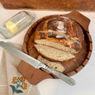 Loaf of bread with a knife on a wooden board, with a decorative napkin and butter dish in the background.
