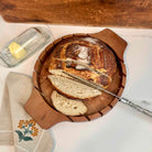 Sliced bread on a wooden board with a knife, butter, and a towel on a marble surface.
