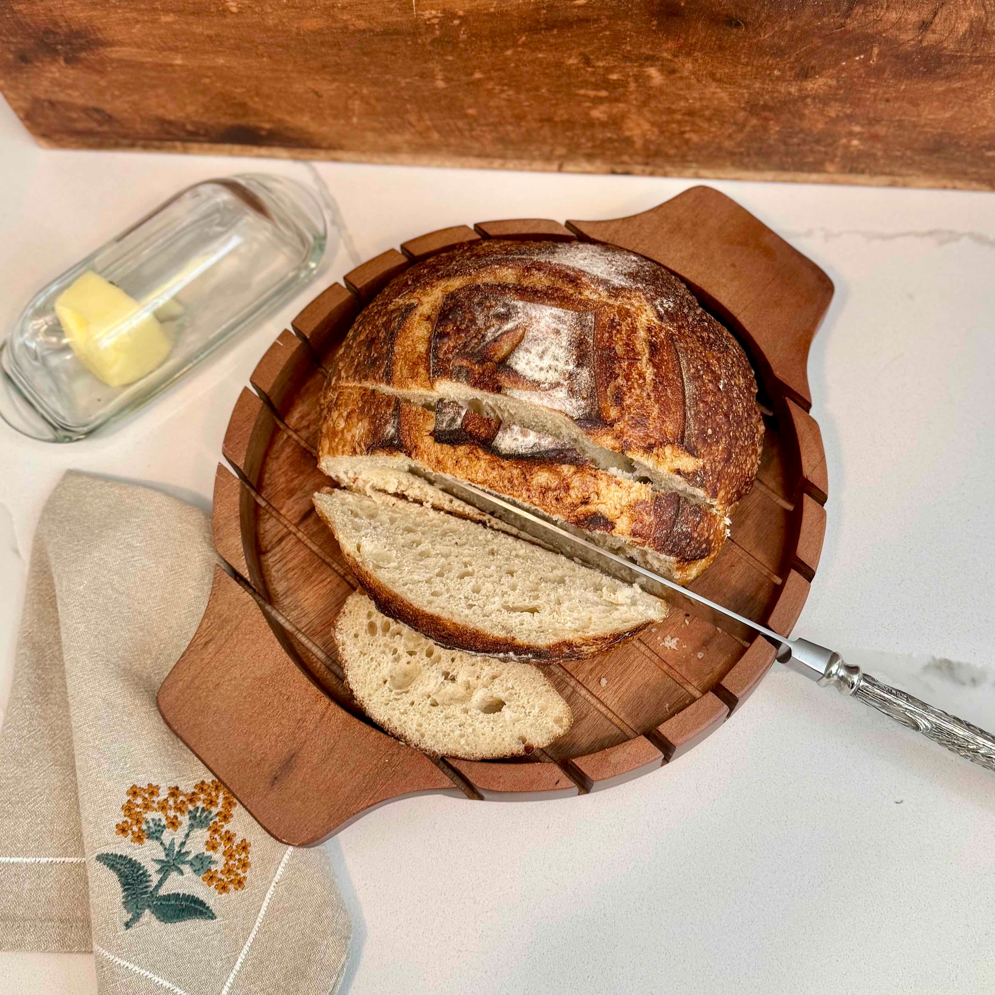 Sliced bread on a wooden board with a knife, butter, and a towel on a marble surface.