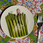 A white plate of cooked asparagus spears sprinkled with grated cheese, placed on a colorful floral tablecloth next to a fork.