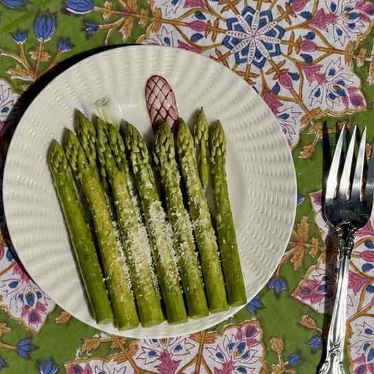 A white plate of cooked asparagus spears sprinkled with grated cheese, placed on a colorful floral tablecloth next to a fork.