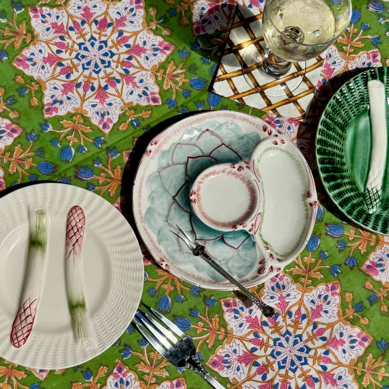 A styled tabletop setting featuring a green tablecloth with a colorful floral mandala pattern, a central ceramic plate with a matching small bowl, and other coordinating plates and cutlery.