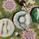 A styled tabletop setting featuring a green tablecloth with a colorful floral mandala pattern, a central ceramic plate with a matching small bowl, and other coordinating plates and cutlery.