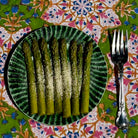 A plate of cooked asparagus spears sprinkled with grated cheese, served on a green patterned plate next to a silver fork on a colorful floral tablecloth.
