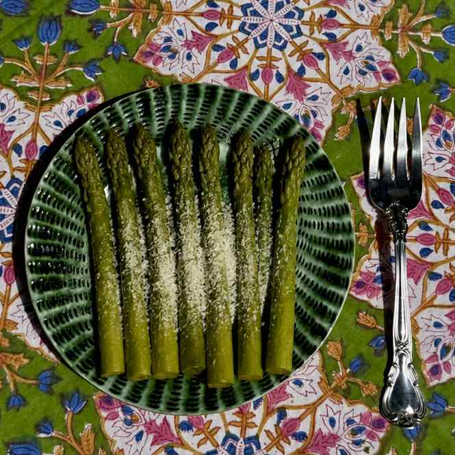 A plate of cooked asparagus spears sprinkled with grated cheese, served on a green patterned plate next to a silver fork on a colorful floral tablecloth.