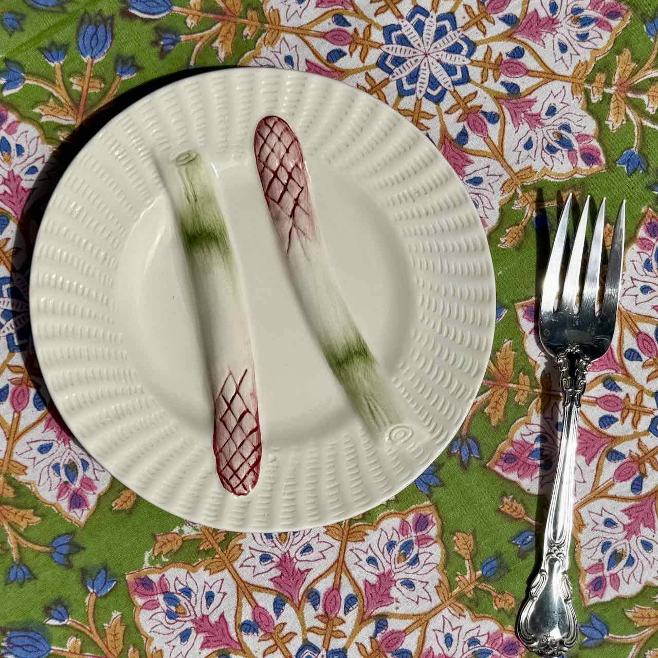 A white ceramic plate with a textured rim and printed asparagus motifs, shown on a colorful floral tablecloth with a silver fork beside it.