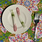 A white ceramic plate with a textured rim and printed asparagus motifs, shown on a colorful floral tablecloth with a silver fork beside it.