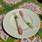A white ceramic plate with a textured, ribbed rim and a raised center medallion, shown on a colorful floral tablecloth. Two decorative leek-shaped objects with red and green patterns are placed on top of the plate.