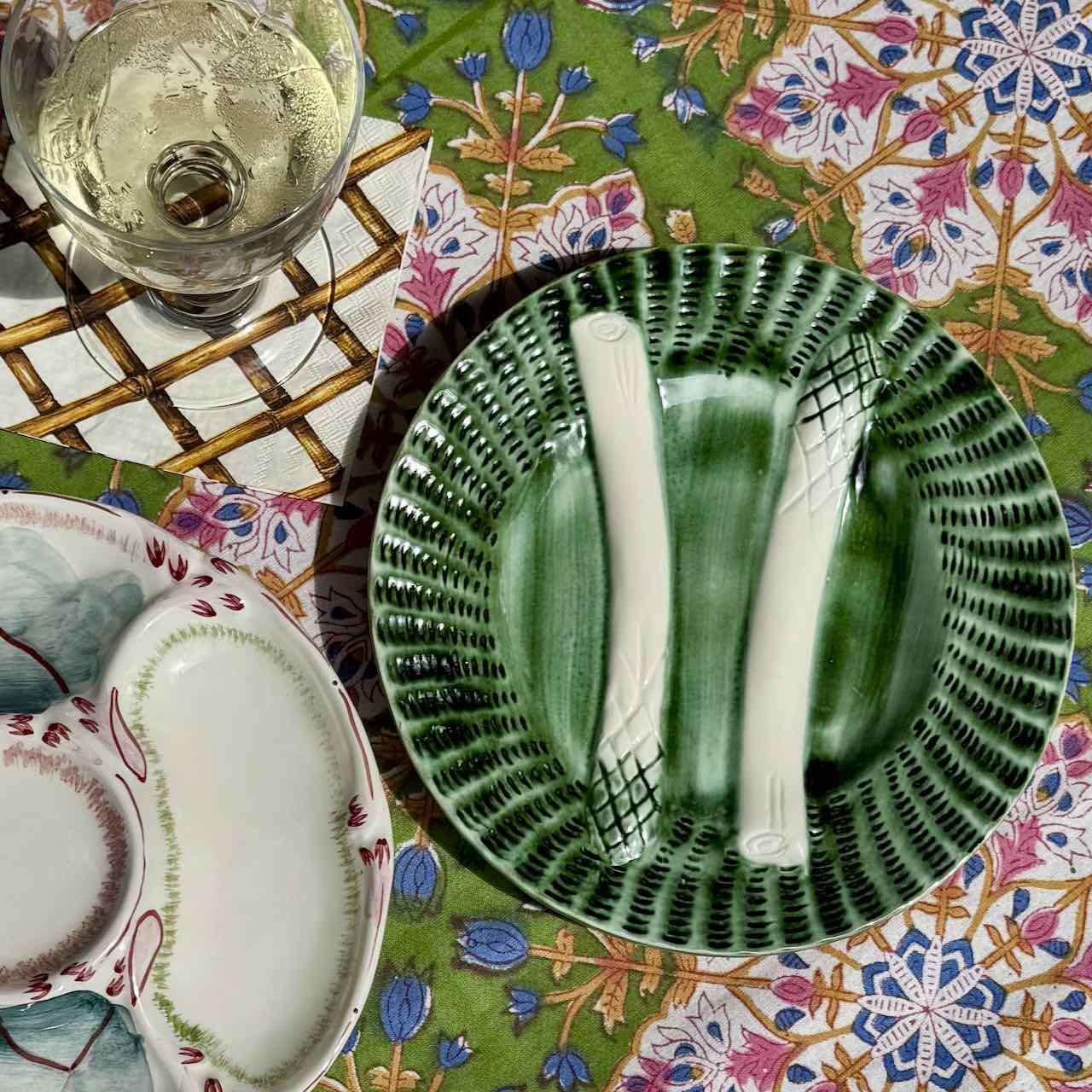 A green ceramic plate with a textured, ribbed rim and two white asparagus spears, styled on a colorful floral tablecloth next to a glass of white wine.